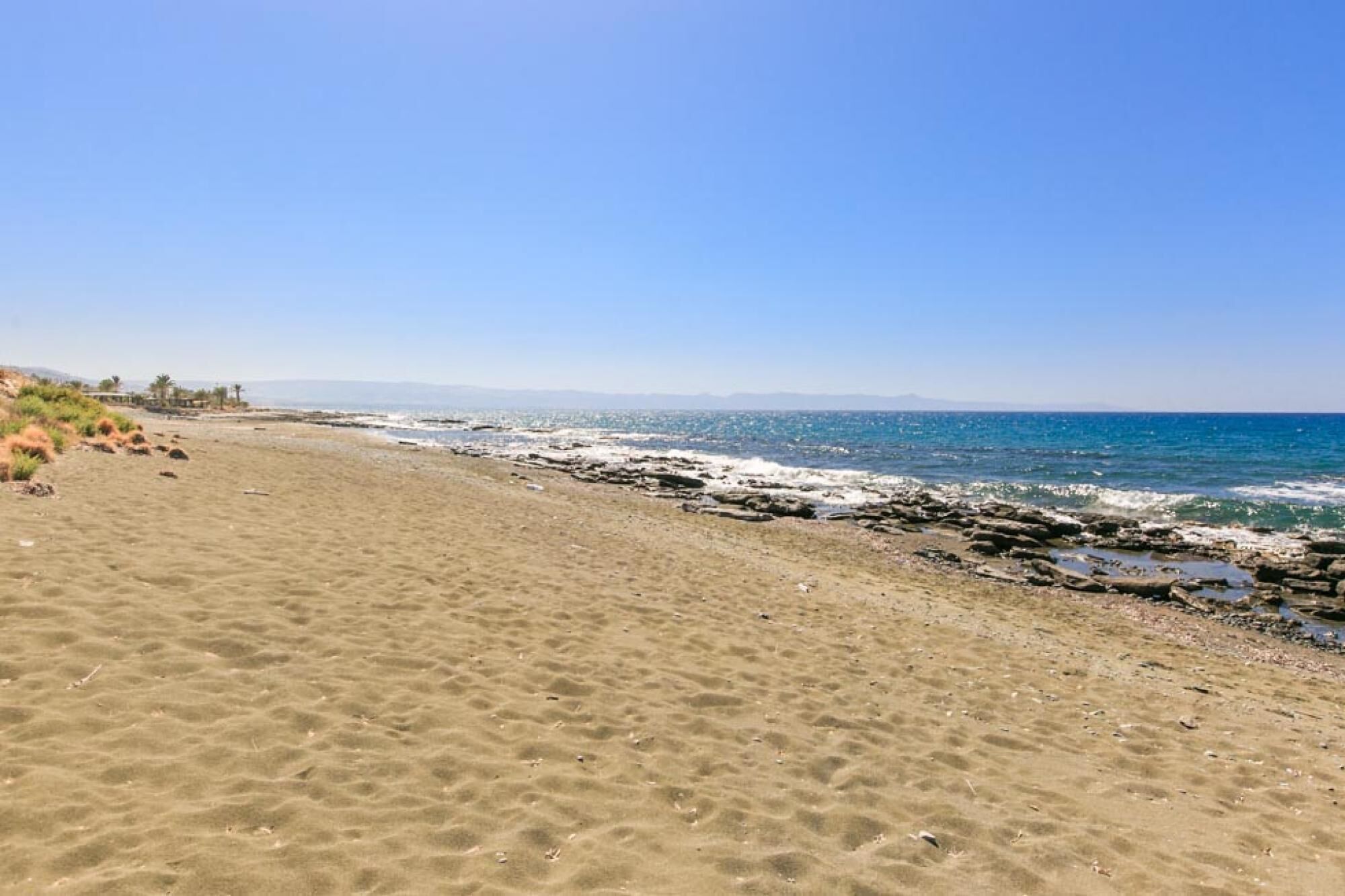 Uma praia arenosa com afloramentos rochosos e um céu azul límpido.