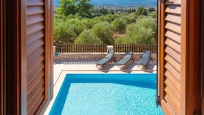 An open wooden door revealing a swimming pool with two lounge chairs and a view of a mountainous landscape.