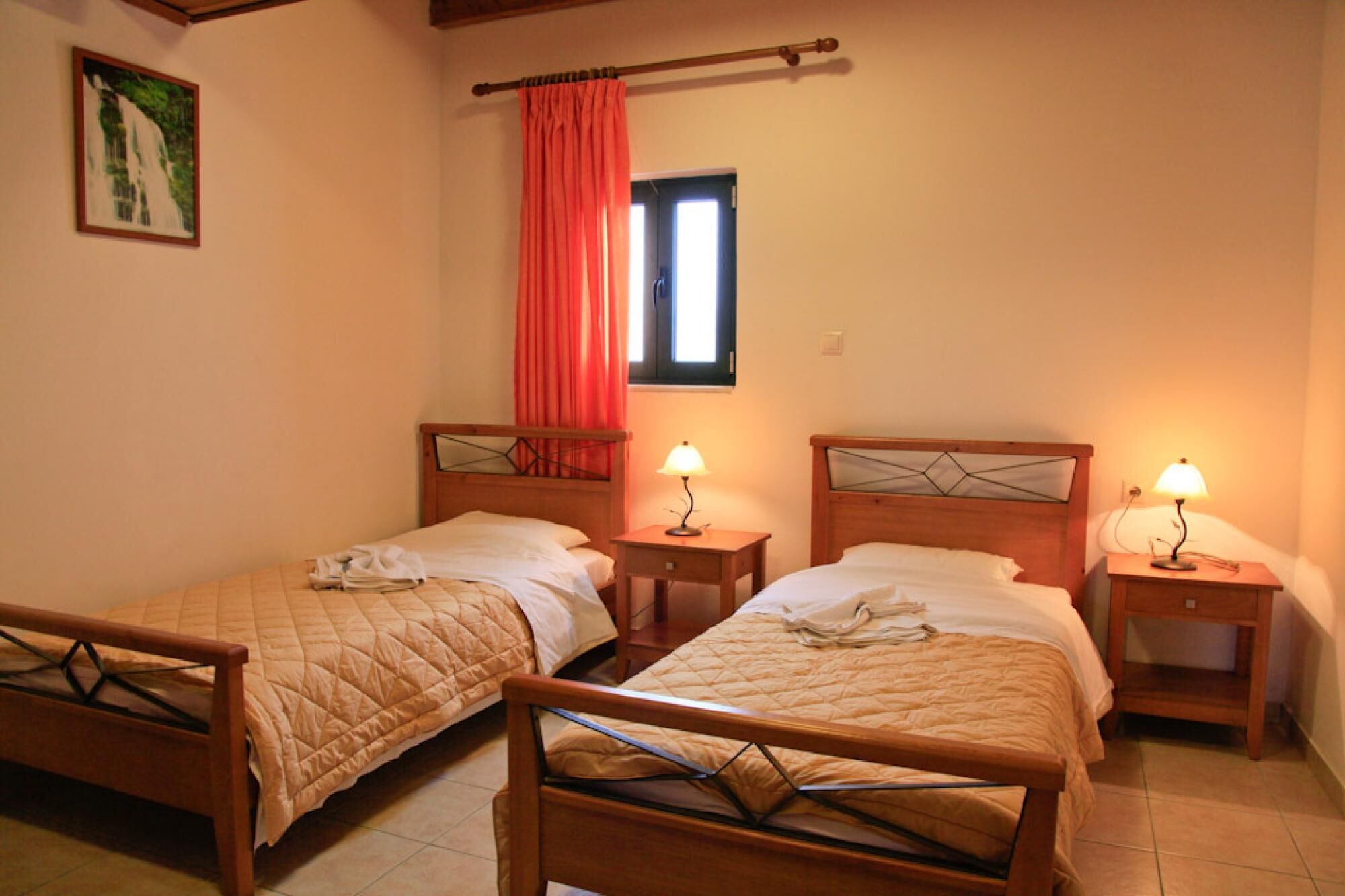 Una habitación con dos camas de madera, cada una con una mesita de noche y una lámpara. Hay una ventana con cortina roja y un cuadro en la pared.
