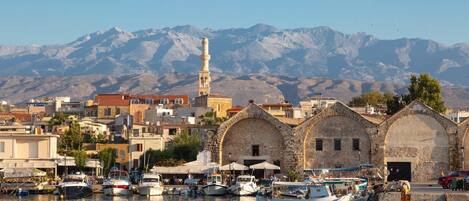 A coastal town with a prominent mosque and three stone buildings with arches. Boats are docked along the waterfront.