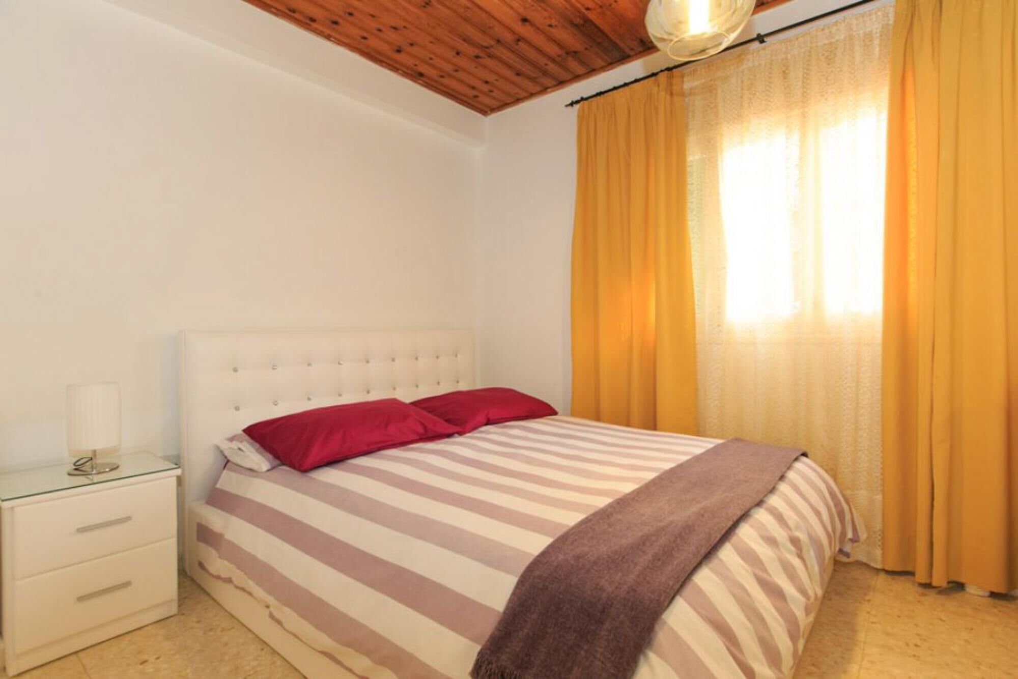 Un dormitorio con edredón a rayas, dos almohadas rojas, un plaid marrón, una mesita de noche blanca con cajones y techo de madera.
