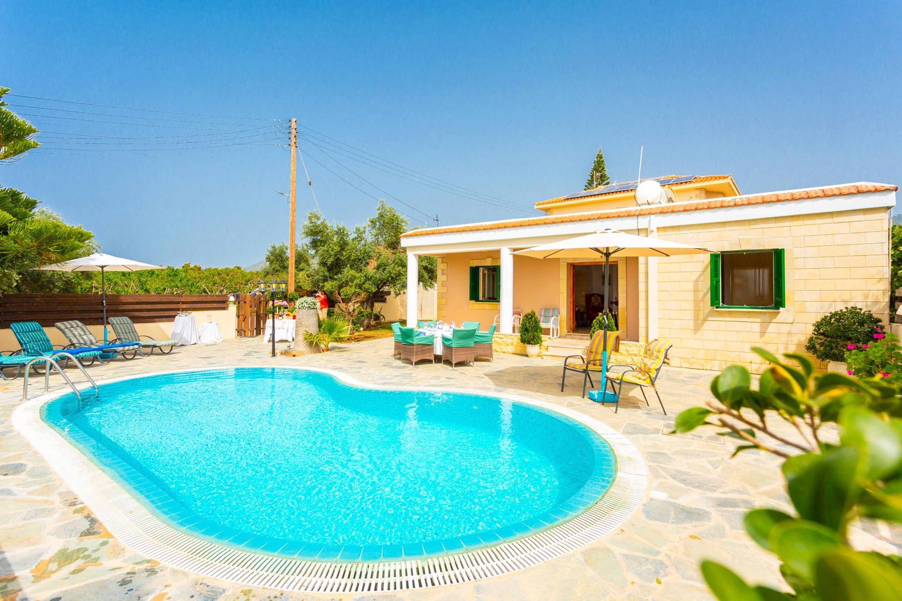Une piscine entourée de chaises longues et d’un espace patio, le tout bordé par une maison avec un toit en tuiles et une porte dotée d’un cadre vert.