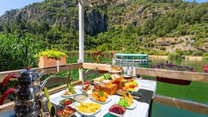 A table set with various dishes and drinks on a deck overlooking a lake, with a boat and forested hills in the background. - Villa Emel Paradise Private Pool A C Wifi Car Not Required Eco-friendly - 2232 (Ortaca)