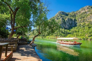 A wooden deck with tables and benches overlooking a river, with a boat passing by and a mountainous backdrop.