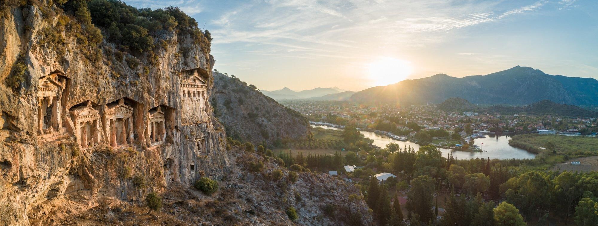 Un penya-segada amb antigues construccions excavades a la roca, un riu i una vila al fons.