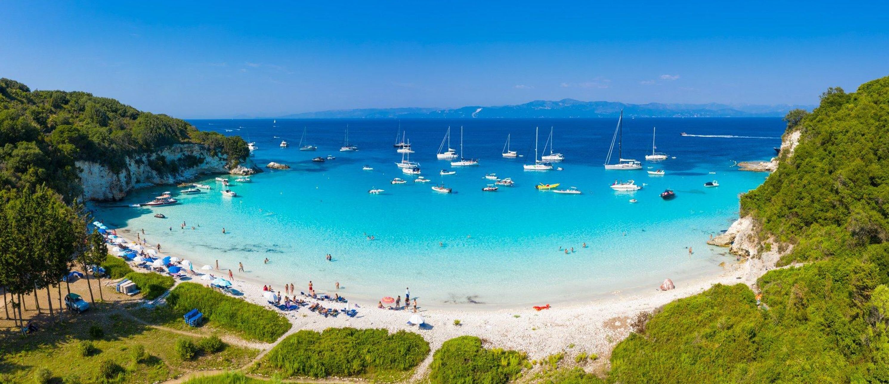 A coastal beach with clear turquoise waters, numerous sailboats, and a sandy beach area with umbrellas and people.