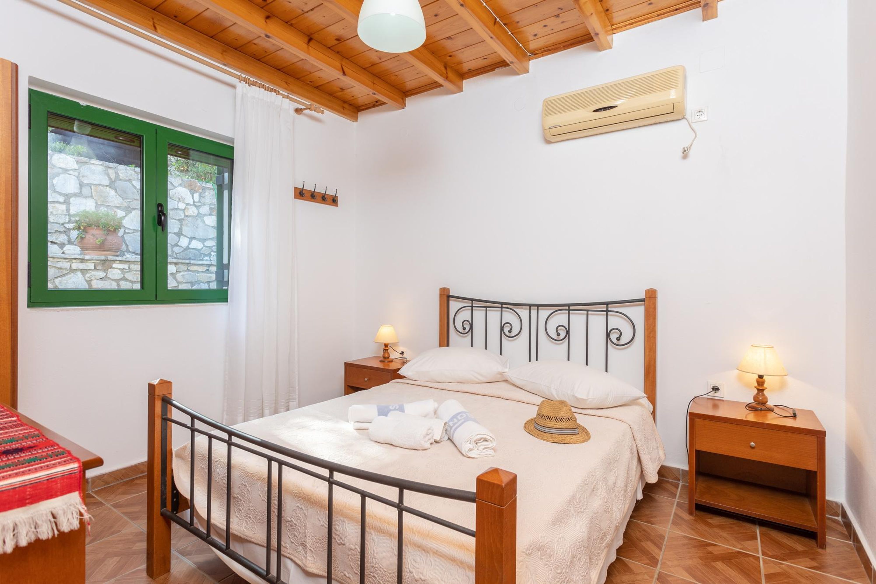 A bedroom with a wooden bed, two bedside tables with lamps, a window with a stone wall view, and a ceiling fan.
