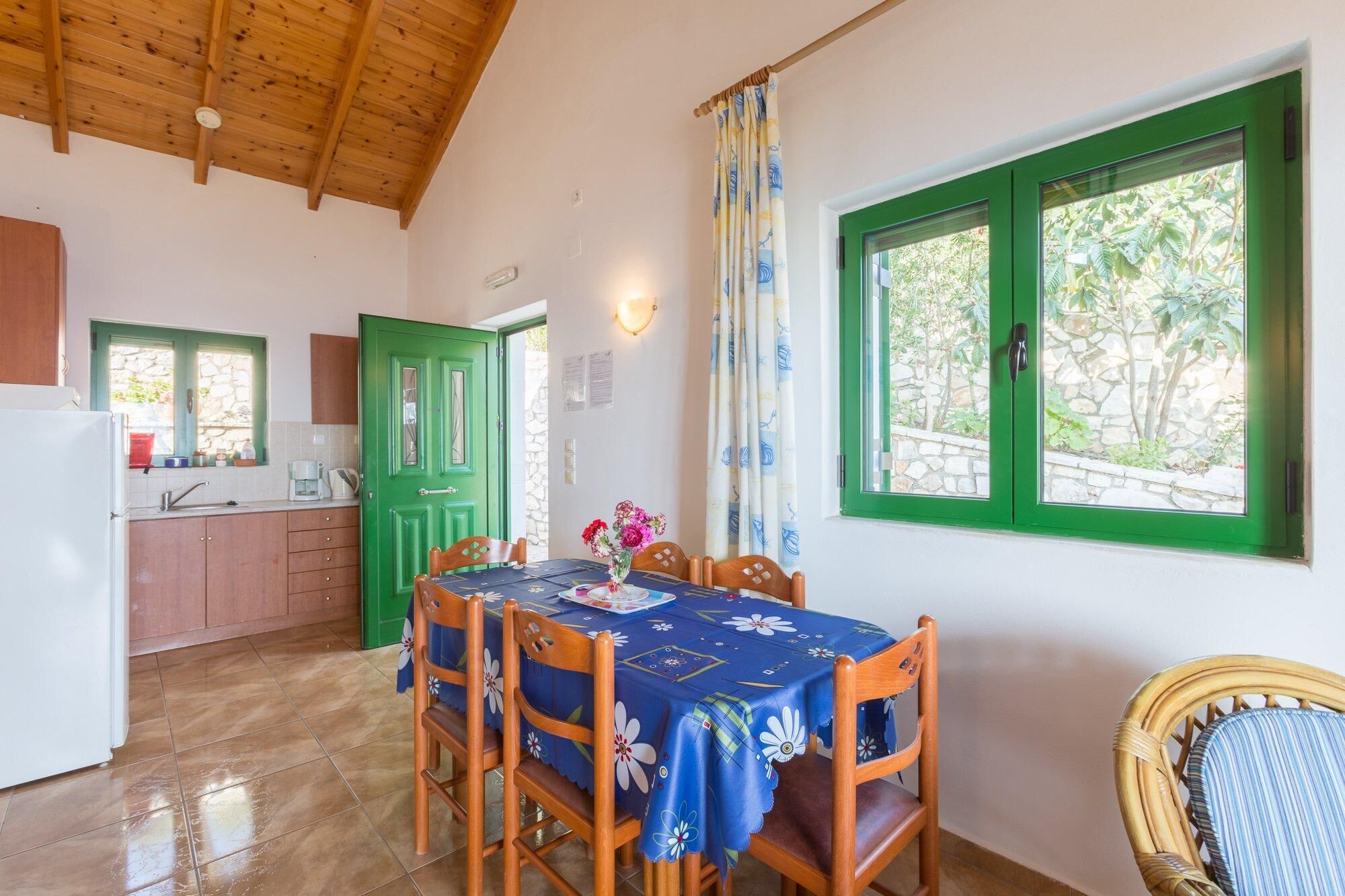 A dining area with a table set for four, wooden chairs, a green door, and a window with a view of greenery.