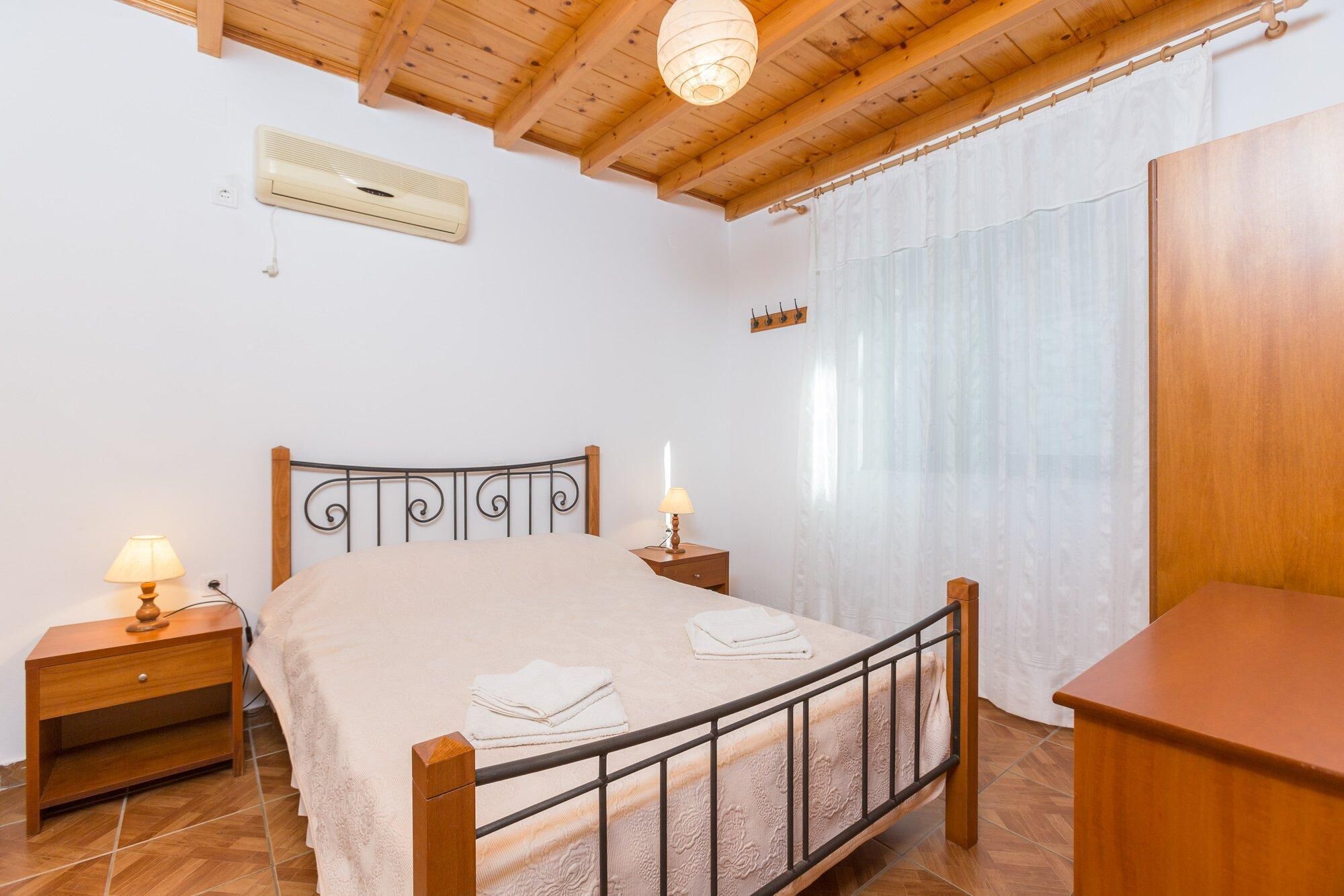 Un dormitorio con techo de madera, una cama con estructura metálica, dos mesitas de noche con lámparas, un revistero de madera, un aire acondicionado instalado en la pared y una ventana con cortinas translúcidas.