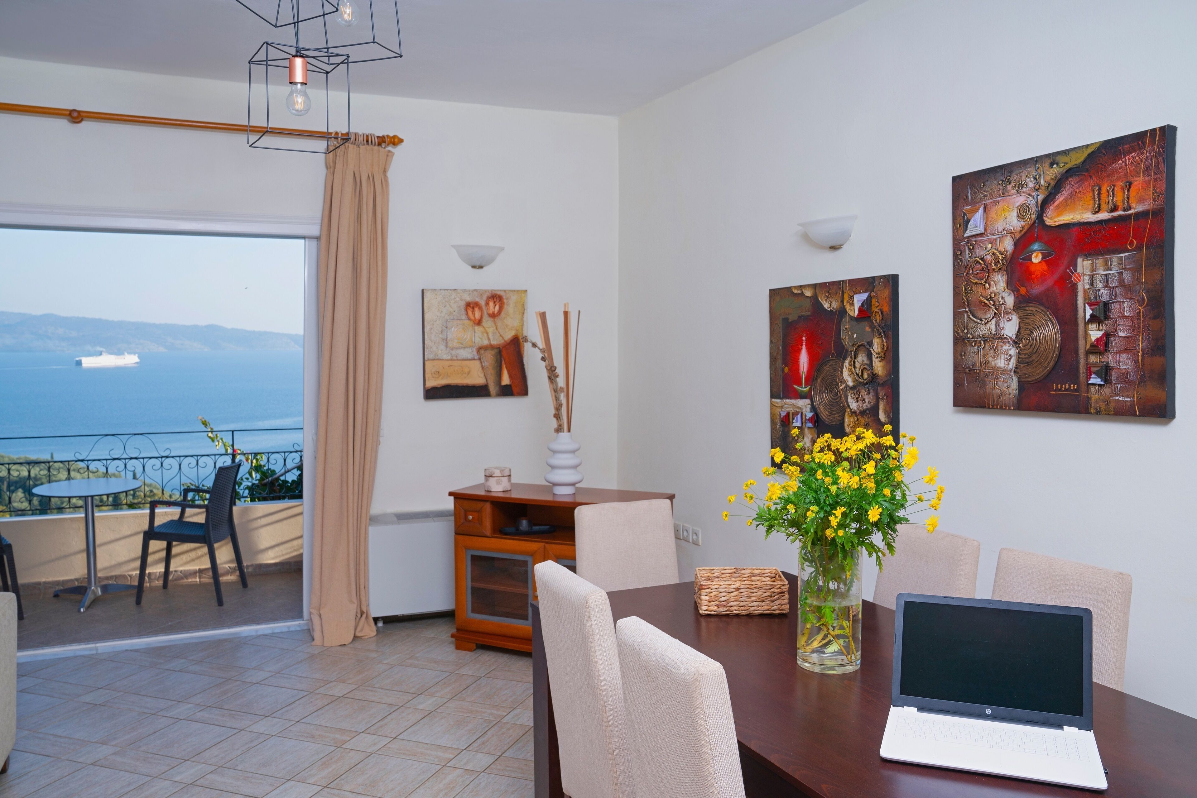 Un área de comedor con una computadora portátil sobre una mesa, un jarrón con flores amarillas y obras de arte en las paredes.