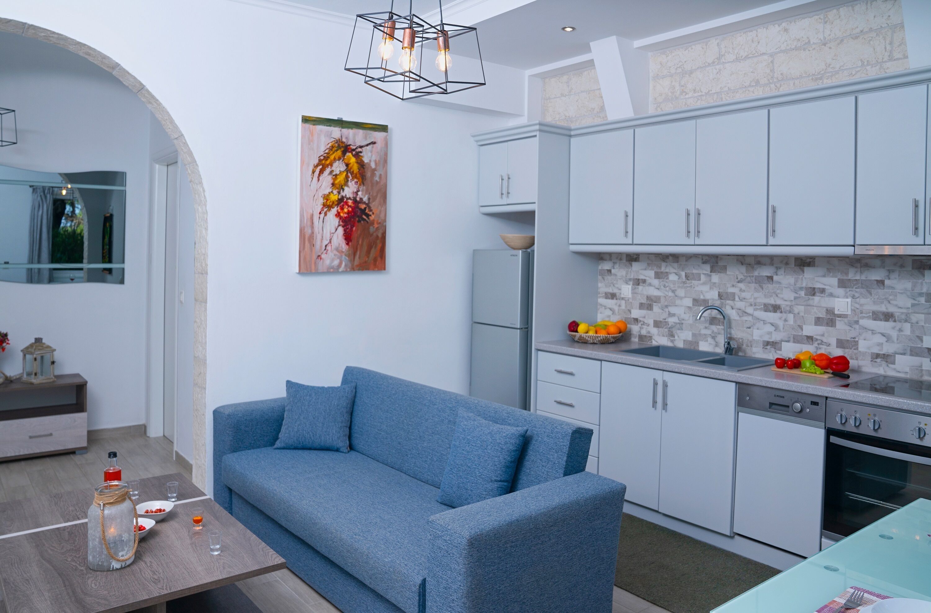Eine moderne Küche mit weißen Schränken, einem eingebauten Ofen und einer Spüle. Im Wohnbereich befindet sich ein blaues Sofa und ein Esstisch mit einer Vase und einer Flasche.