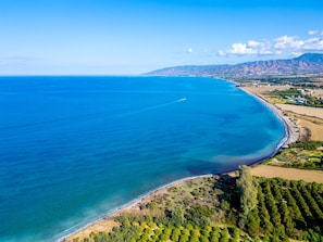 Vista aérea de una zona costera con mar azul cristalino, una playa de arena y un paisaje montañoso al fondo.