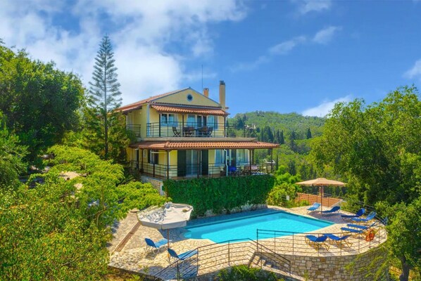 Una casa de dos pisos con piscina, rodeada de vegetación exuberante y colinas.