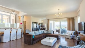 Un vaste salon avec un canapé d’angle marron, une table basse blanche et un coin repas avec des chaises à rayures. On aperçoit une cuisine au fond, et il y a un balcon avec vue.