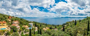 A coastal village with houses, a swimming pool, and a view of the sea and distant mountains.
