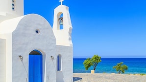 Un edificio blanco con puerta azul y un campanario, con vista al mar azul y despejado.