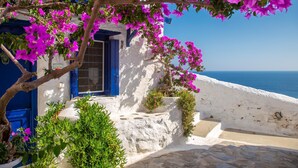 Un edificio blanco con contraventanas azules y flores de buganvilla rosadas, un área empaveda de piedra y vistas al mar.