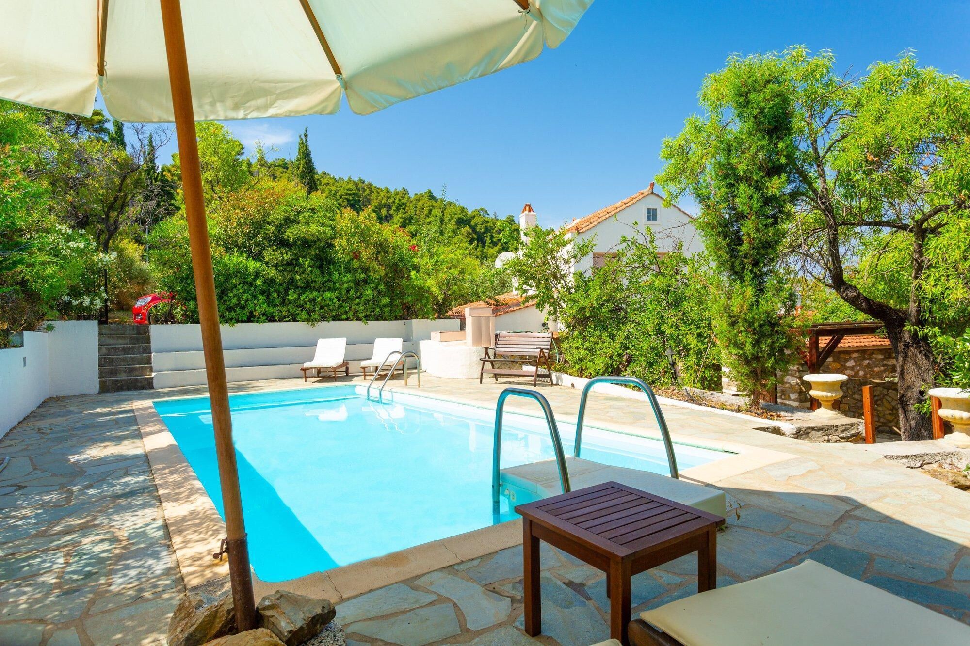 A swimming pool with lounge chairs and a table, surrounded by stone tiles and greenery.