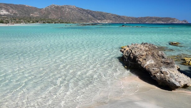Un mar turquesa transparente con un promontorio rocoso en primer plano y montañas al fondo.