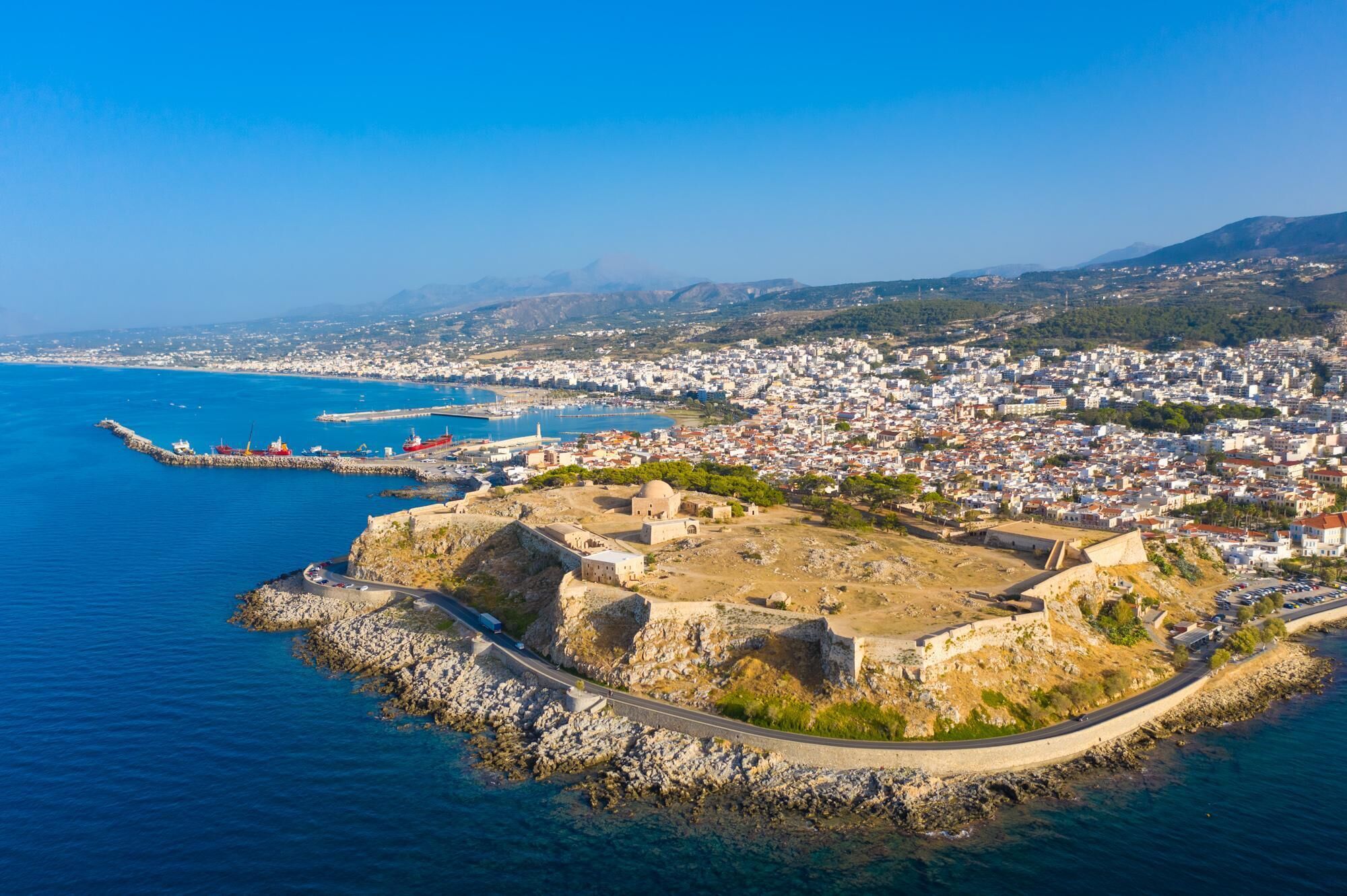 Vue aérienne d’une ville côtière, avec une imposante fortification historique sur une péninsule rocheuse, un port avec des navires, et une zone urbaine densément peuplée en arrière-plan.