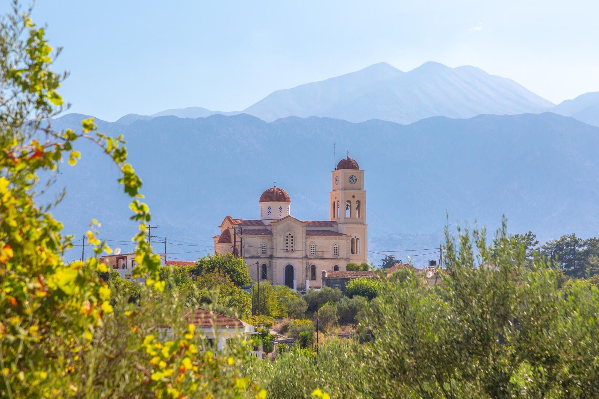 A church with two domes and a bell tower, surrounded by greenery and mountains in the background.