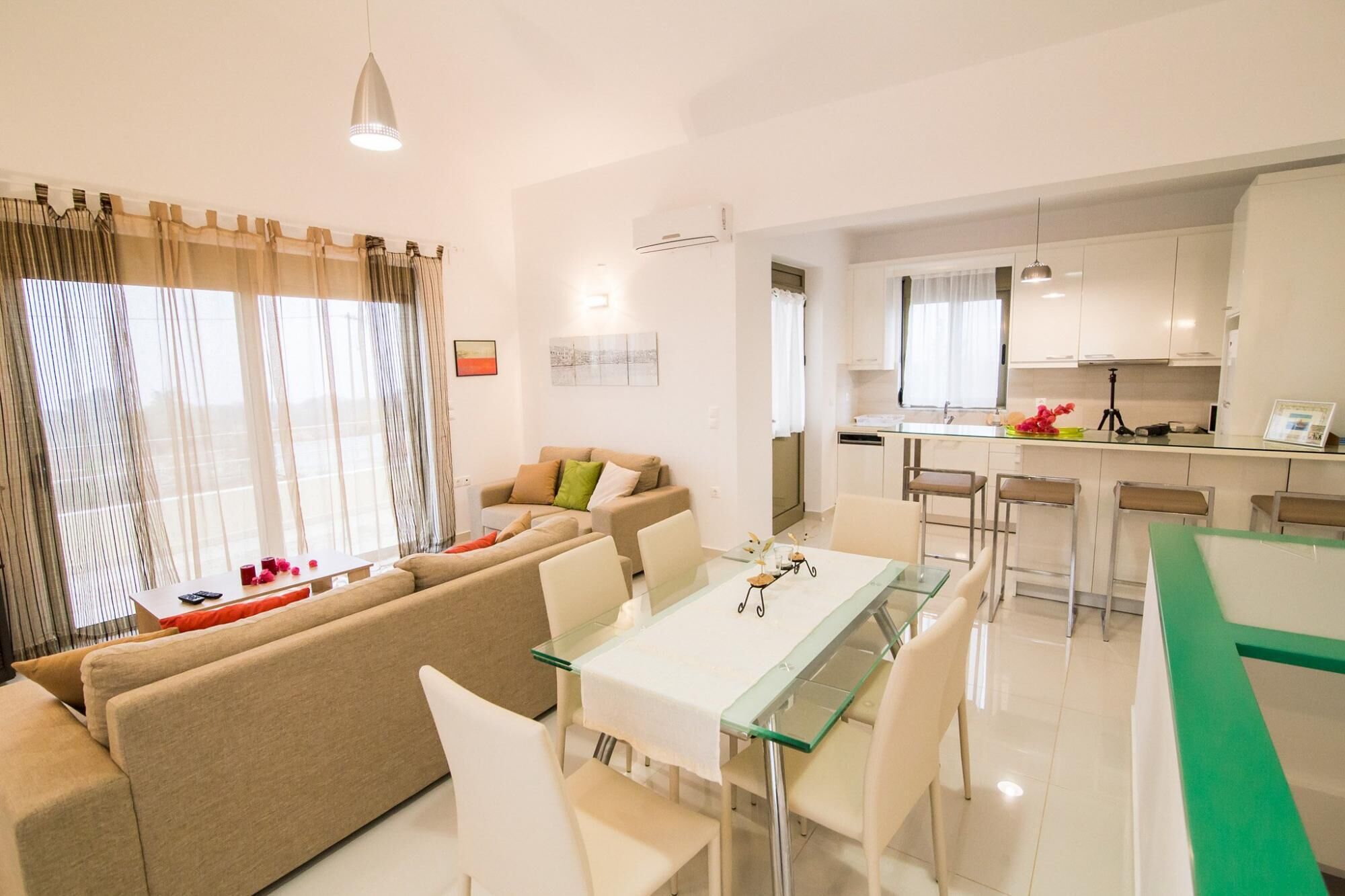 A modern living room with a glass dining table, a sofa, and a kitchen area with white cabinets and a bar counter.