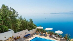 Une piscine rectangulaire bordée de pierres, entourée de chaises longues et de parasols, d’où l’on peut admirer une mer d’un bleu limpide.