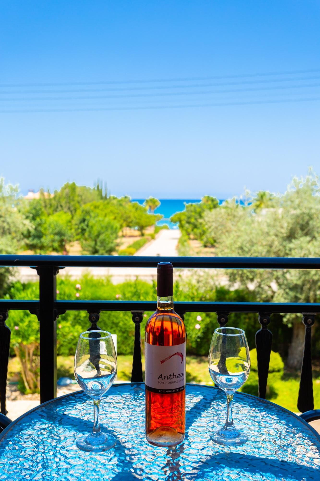 A table with a bottle of wine and two glasses on a balcony overlooking a garden and a path leading to a body of water.