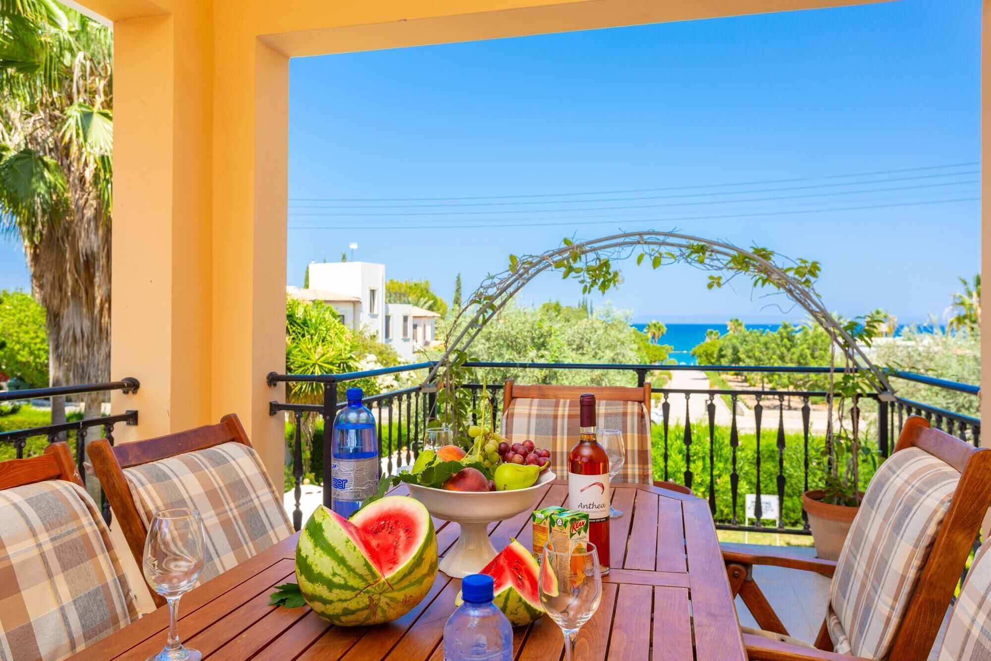 A balcony with a table set for a meal, featuring a bowl of fruit, a watermelon, and a bottle of juice.