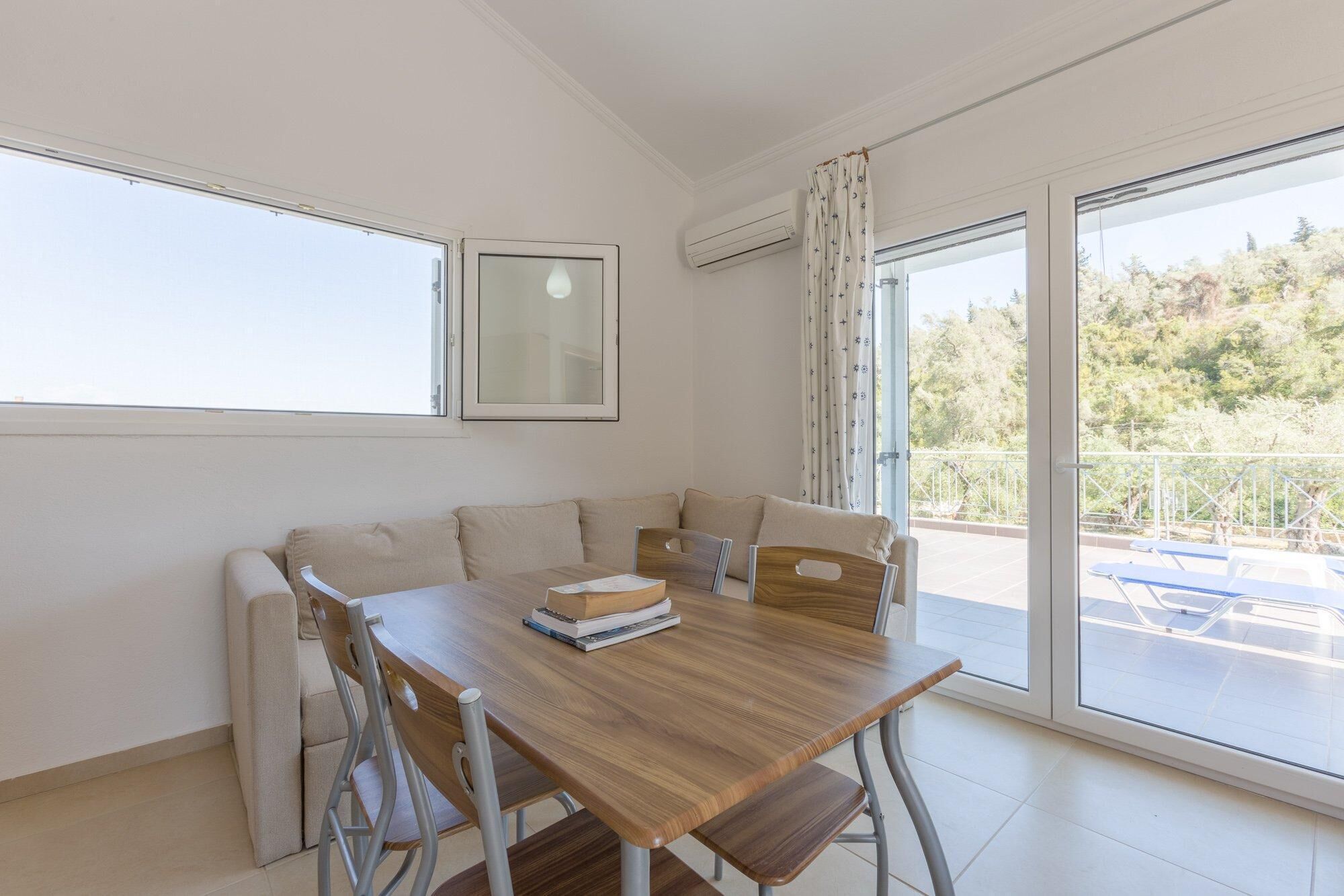 A modern room with a wooden dining table, beige sofa, and a balcony with a view of trees.