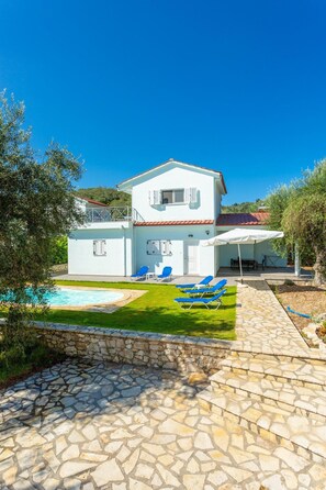 Une maison blanche à deux étages, dotée d’une piscine, de chaises longues bleues et d’une terrasse en pierre.