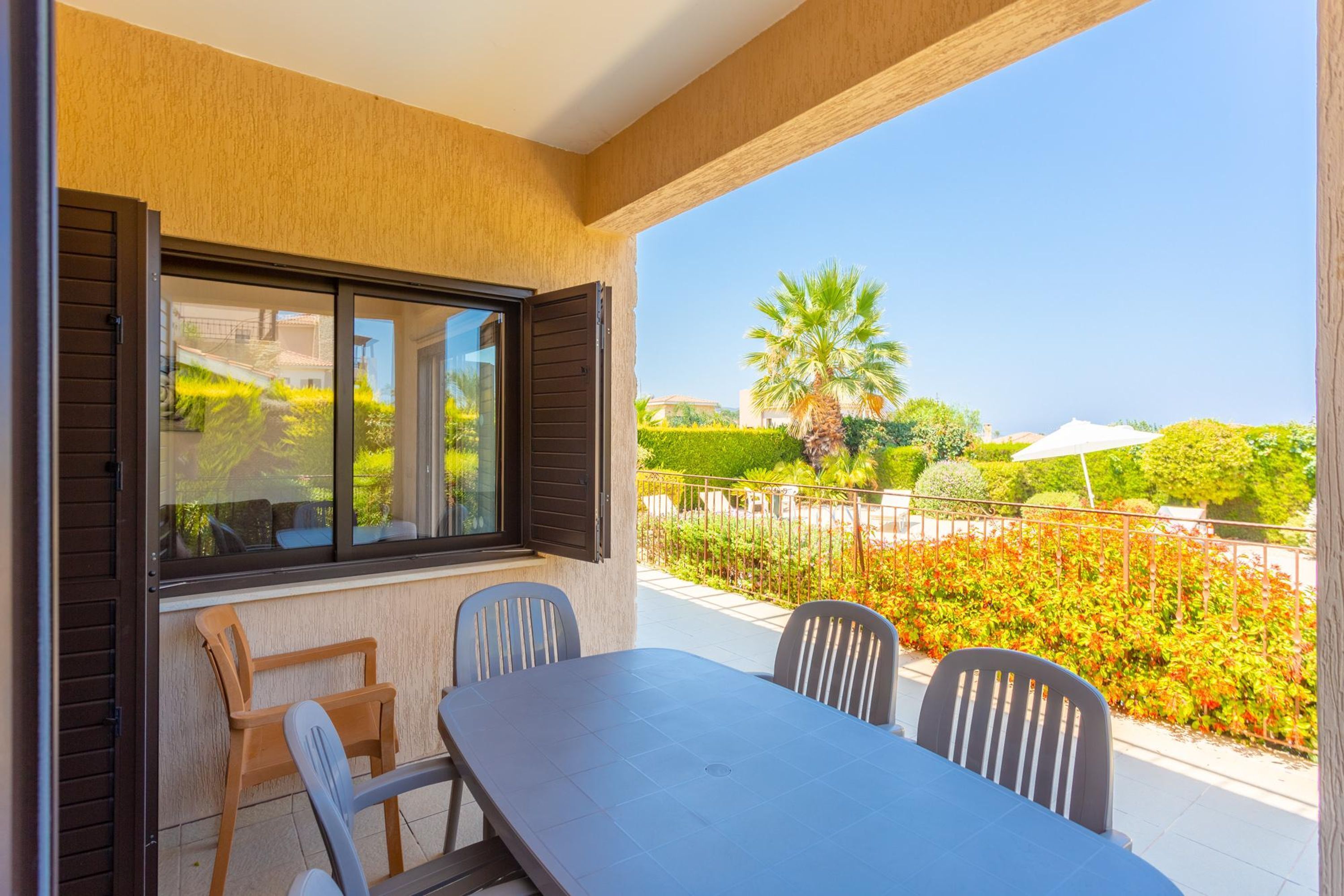 An outdoor patio with a table and chairs, a view of a landscaped garden, and a clear blue sky.