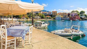 A seaside restaurant with outdoor seating, a view of colorful buildings, and boats in the water. - Villa Erasmia Large Private Pool Walk to Beach Sea Views A C Wifi - 2646 (Kefalonia)