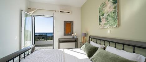 Une chambre avec un grand lit, une vue sur la mer par une porte ouverte, une petite table de chevet avec une lampe et un tableau accroché au mur.