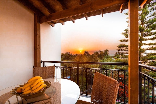 A balcony with a table and chairs, a bowl of fruit, and a view of the sunset.