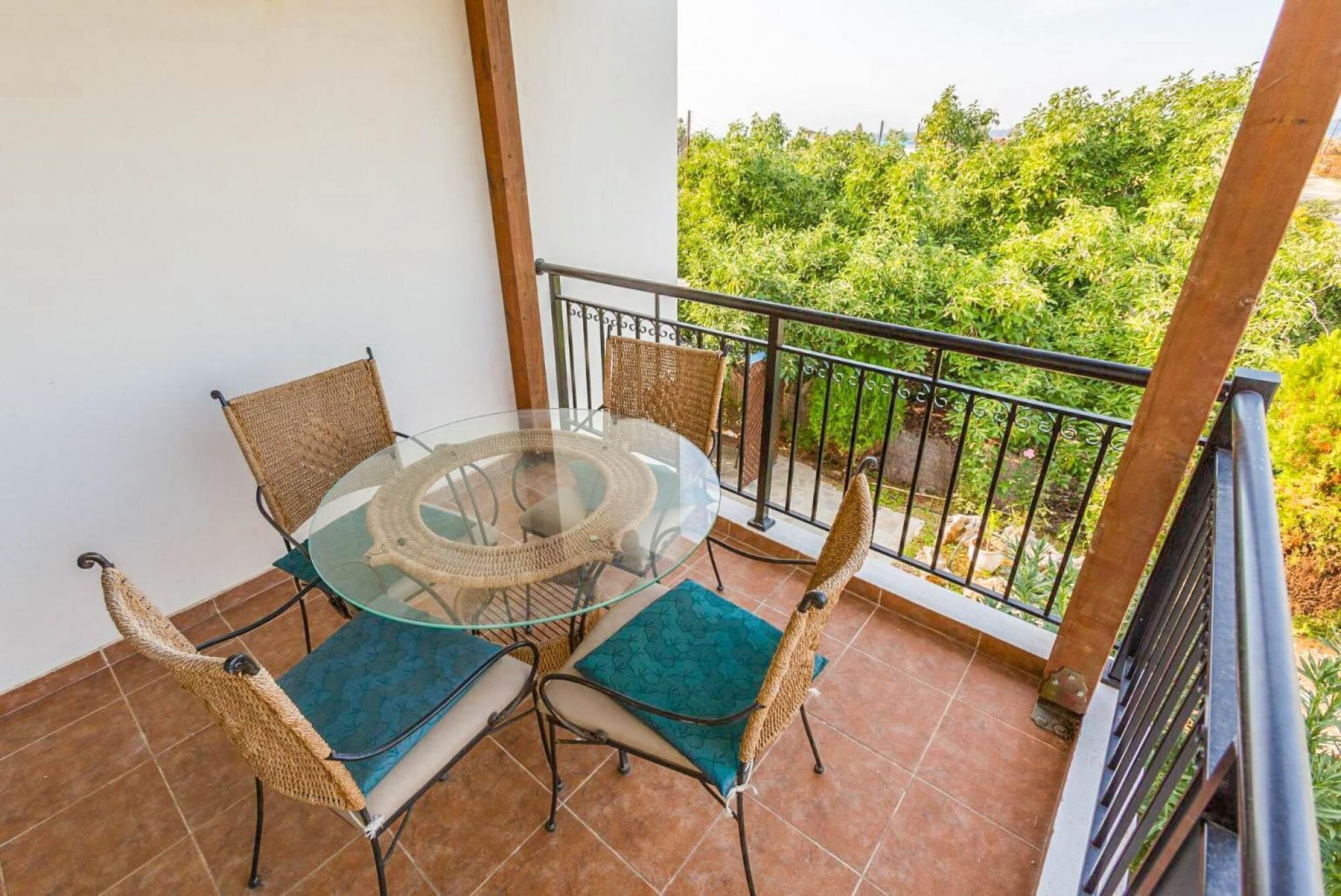 A balcony with a glass table and wicker chairs, overlooking a lush garden.