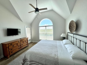 Un dormitorio con cama, cómoda, televisor, ventilador de techo y ventana con persianas.