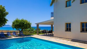 A swimming pool with lounge chairs and a table, a white building with windows, and a view of the sea.