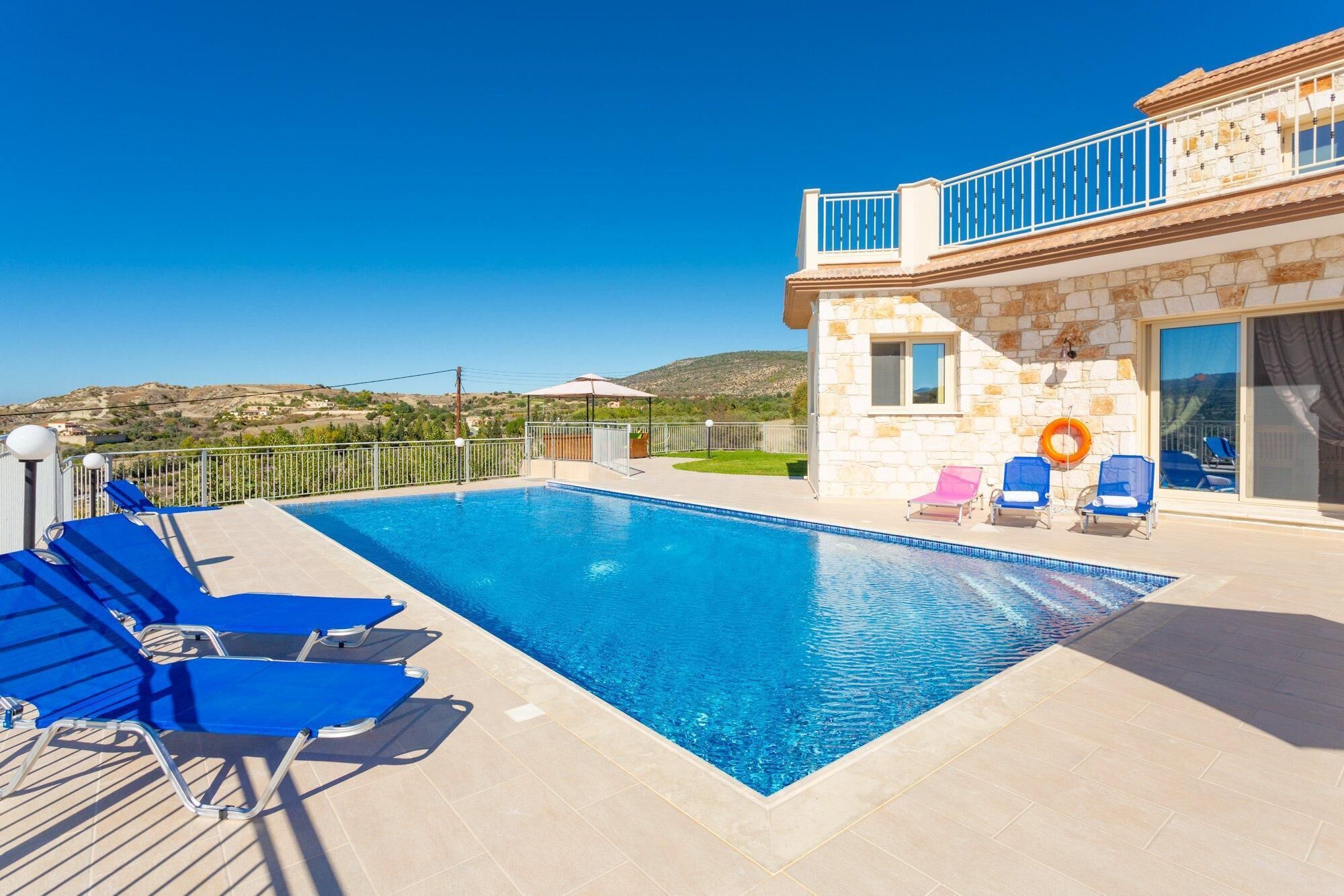 A swimming pool with blue lounge chairs, a stone building with a balcony, and a clear view of the hills in the background.