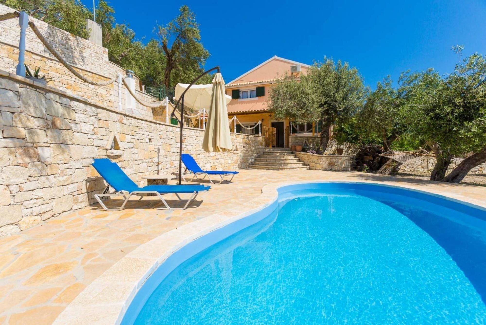 A pool area with stone walls, a house in the background, and a clear blue sky.