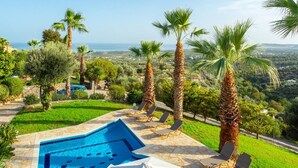 Piscina con vista al mar, rodeada de palmeras y un patio de piedra con mobiliario exterior.
