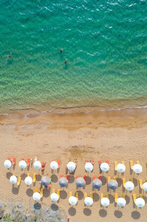 Vue aérienne d’une plage aux eaux turquoise et limpides, avec un rivage sablonneux et de nombreuses rangées de parasols et de chaises de plage.