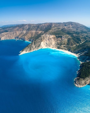 Une vue aérienne d’une zone côtière, avec une mer bleue et limpide, une plage de sable et un relief accidenté.