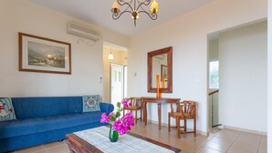A living room with a blue sofa, a wooden coffee table with a checkered tablecloth, a vase with pink flowers, a wooden dining table with chairs, a mirror, and an air conditioning unit on the wall. - Skala Villa Green Large Private Pool Walk to Beach Sea Views A C Wifi - 2825 (Kefalonia)