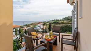Un balcon agrémenté de meubles en osier, une table dressée avec de la nourriture et des boissons, et une vue sur la mer et les collines.
