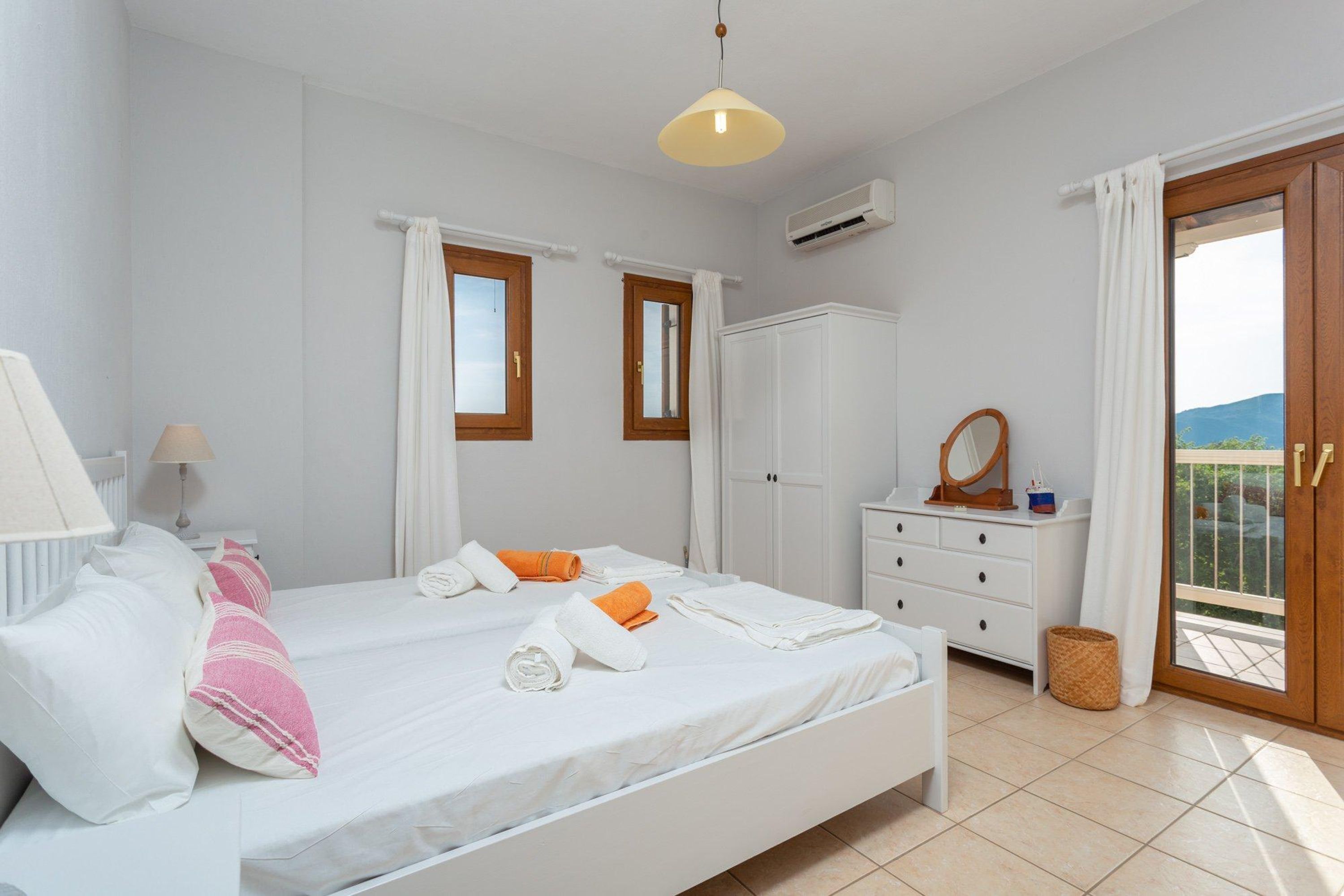 Een slaapkamer met een bed, witte muren, een raam met gordijnen, een dressoir en een balkondeur.