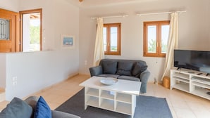 A modern living room with a grey sofa, a white coffee table, a TV stand, and large windows with curtains. - Villa Glafki Large Private Pool Sea Views A C Wifi - 2829 (Skopelos)