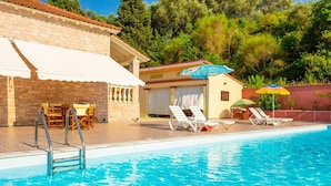 Une piscine avec des chaises longues et des parasols, un bâtiment en pierre avec un toit en tuiles, et une vue sur les arbres en arrière-plan.