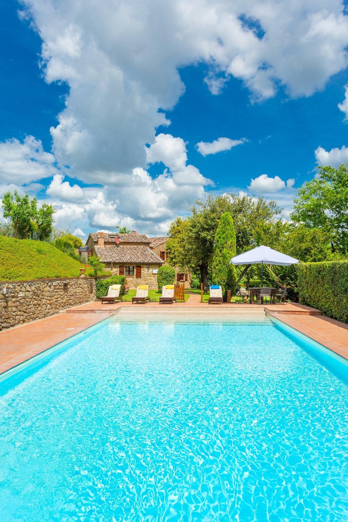 A swimming pool with lounge chairs and umbrellas, surrounded by a stone wall and greenery.