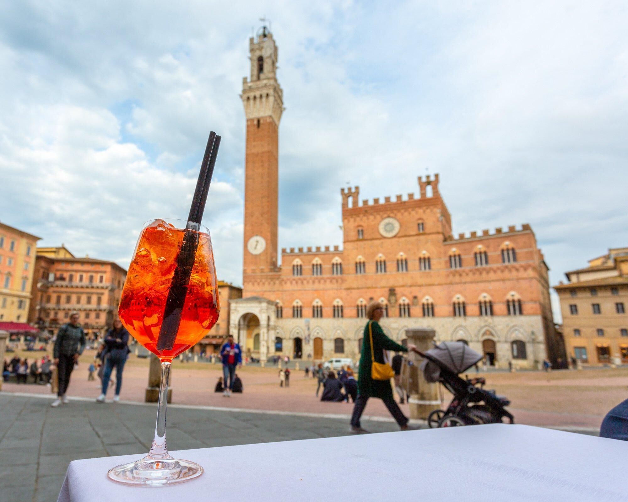 A glass of red drink with a straw on a table in a public square with historic buildings in the background.